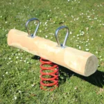 Foto der Federwippe Duo aus einem dicken Robinienstamm mit zwei gegenüberliegenden Edelstahlgriffen; dicke, stabile Schwingfeder unterhalb des Sitzstamms, in der Mitte zwischen den Griffen angebracht. Hintergrund: saftig-grüne Frühlingswiese mit Gänseblümchen