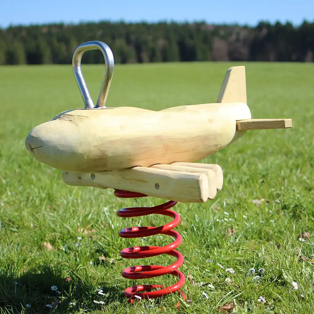 Federwippe Flugzeug aus Robinienholz mit Tragflächen aus dünnen Stammabschnitten sowie Heckruder; auf einer roten, kräftigen Schwingfeder; Edelstahlgriff; Spielgerät steht auf einer grünen, saftigen Frühjahrswiese mit Gänseblümchen; Im Hintergrund in der Entfernung eine dunkle Waldkante vor strahlend blauem Himmel