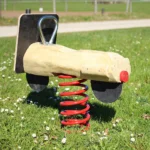 Foto der Federwippe Retro-Roller Ansicht von schräg hinten mit Blick auf Rückseite. Dort Rückleuchgte erkennbar. Hintergrund: saftig-grüne Frühlingswiese mit Gänseblümchen dahinter Kiesweg