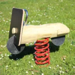 Foto der Federwippe Retro-Roller aus einem dicken Robinienstamm mit Edelstahlgriff; dicke, stabile Schwingfeder unterhalb des Sitzstamms, Reifen aus Gummimatten angedeutet; kleine Details wie Frontleuchte, Windschutzschild etc. Hintergrund: saftig-grüne Frühlingswiese mit Gänseblümchen