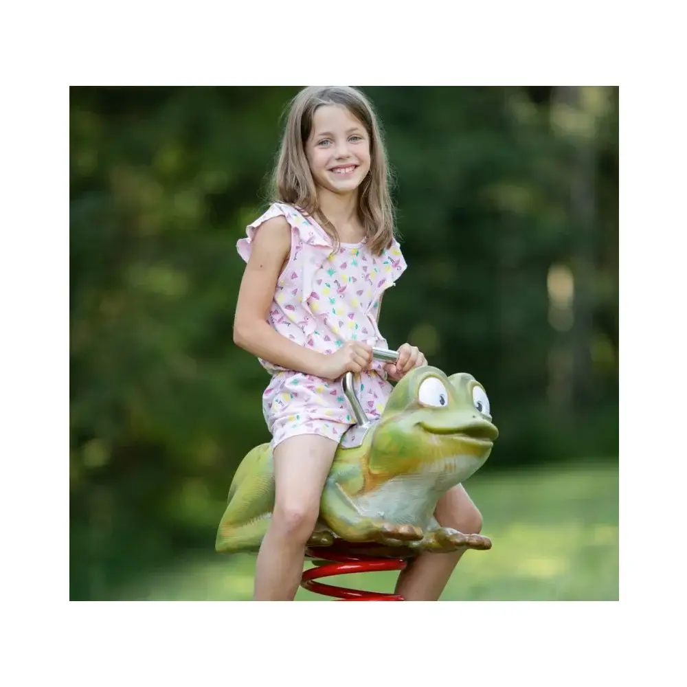 Foto: geschnitzes und bemaltes Federwipptier grüner Frosch. Mädchen sitzt auf seinem Rücken; unscharfer, dunkelgrüner Hintergrund