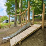 Podest für Hangrutsche mit Aufstiegsrampe im Wald; direkt daneben eine Edelstahl-Hangrutsche. Im Hintergrund verläuft ein Motorikpfad aus Robinienstämmen mit verschiedenen Spielelementen