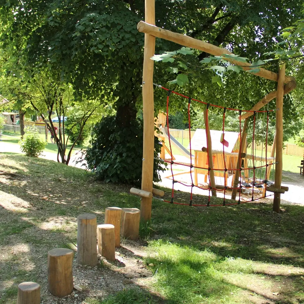 Motrikpfad für Kindergartenkinder unter einem Baum an einem leichten Hang. Im Vordergrund: Balanicerpoller, dann ein waagrechtes Kletternetz, Im Hintergrund ein Spielhaus.