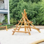 Klettergerüst Pyramide auf Fallschutzsand-Fläche. Im Hintergrund eine grüne Laubhecke und angeschnitten die Ecke eines Wohnhauses. Teil von Wohnquartier Spielplätze
