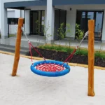 Mininestschaukel-Robinie Eine Mini-Nestschaukel mit rotem Netz und blauer Umrandung, niedrig aufgehängt an zwei Robinienpfosten, steht auf weißem Sand. Im Hintergrund eine leichte, begrünte Böschung zu einer Terrasse einer Wohnung