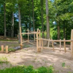 Motorikpfad individuell im Waldstück: Zwischen hohen Buchen schlängelt sich ein Robinien-Abenteuerpfad mit verschiedenen Spiel-, Balance- und Kletterelementen. Etwas Sonnenlicht fällt auf das erste Element im Vordergrund: eine Balancierstrecke aus einer wackeligen Gummimatte