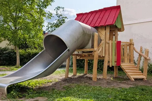 große Edelstahl-Röhrenrutsche angeschlossen an einem erhöhten Spielhaus auf Stelzen aus Robinienholz in einem Innenhof einer Kinderkrippe