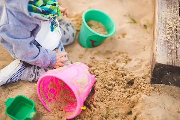 Detailansicht bzw. Ausschnitt: Kind kniet im Sandkasten. Im Spielsand liegen einige Sandspielsachen aus Kunststoff.
Bild illustriert die Füllmenge, so wie im Fallschutz- und Spielsand-Rechner angegeben