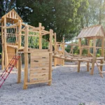Spielturmanlage mit verschieden hohen Türmen aus Robinienholz, zwei davon mit Satteldach, verschiedene Wackelbrücken und Balancierelemente als Verbindungen zwischen den im Quadrat angeordneten Türmen, Fallschutzkies unter den Türmen, im Hintergrund eine dichte Laubhecke mit hohen Ahornbäumen