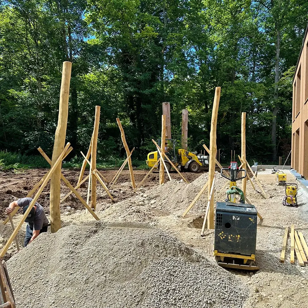 Baustelle neben einem neuen KiTa-Gebäude. In Löchern im Boden stecken Abgestützte Robinienstämme, dazwischen befinden sich einige Kieshaufen des Aushubs. Im Hintergrund ein dunkler Laubwald