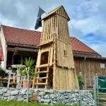 Foto von der Vorderseite des Spielturm Zuckerhut: Der Robinienturm läuft von seiner quadratischen Grundform bis auf ca. 5 m Höhe mehr oder weniger spitz zusammen. die unteren Ebenen sind vollständig verschlagen. die obereste Ebene hat einen lockeren Lattenverschlag mit licht- und blickdurchlässigen Spalten. vom Boden in den ersten STock führt eine Knüppelleiter. über dem Einstieg befindet sich ein kleines Vordach