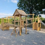 Spielturmanlage mit verschieden hohen Türmen aus Robinienholz, zwei davon mit Satteldach, verschiedene Wackelbrücken und Balancierelemente als Verbindungen zwischen den im Quadrat angeordneten Türmen; Im Vordergrund der niedrigste Turm mit Plattformhöhe 1m und einem Sandaufzug; Fallschutzkies unter den Türmen, im Hintergrund eine dichte Laubhecke und ein hoher Ahornbaum, dahinter ein bewaldter Berg in der Ferne, alles vor blauem Himmel