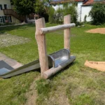 Hangrutsche aus Edelstahl befestigt an einem Startportal aus Robinienstämmen. Blick vom Hügel hinunter über die Rutsche.