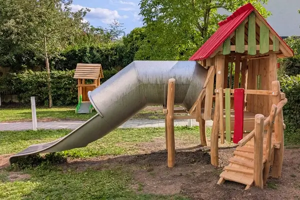 Seitenansicht auf ein Spielhaus mit Zustiegsplattform und kleiner Rampe. nach links aus dem AHus heraus eine Röhrenrutsche