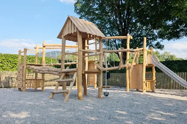 Spielturmanlage mit verschieden hohen Türmen aus Robinienholz, zwei davon mit Satteldach, verschiedene Wackelbrücken und Balancierelemente als Verbindungen zwischen den im Quadrat angeordneten Türmen; Im Vordergrund der niedrigste Turm mit Plattformhöhe 1m und einem Sandaufzug; Fallschutzkies unter den Türmen, im Hintergrund eine dichte Laubhecke und ein hoher Ahornbaum, dahinter ein bewaldter Berg in der Ferne, alles vor blauem Himmel
