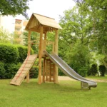 Foto aus Froschperspektive auf viereckigen Spielplatz-Turm aus Robinienholz mit Satteldach, Rampe mit Handlaufseil, Edelstahlrutsche, schräges Kletternetz und Kletterwand. Der Turm steht auf einem gepflegten, kurzgeschnittenen Rasen in einer GRünanlage zwischen hohen Wohnblöcken